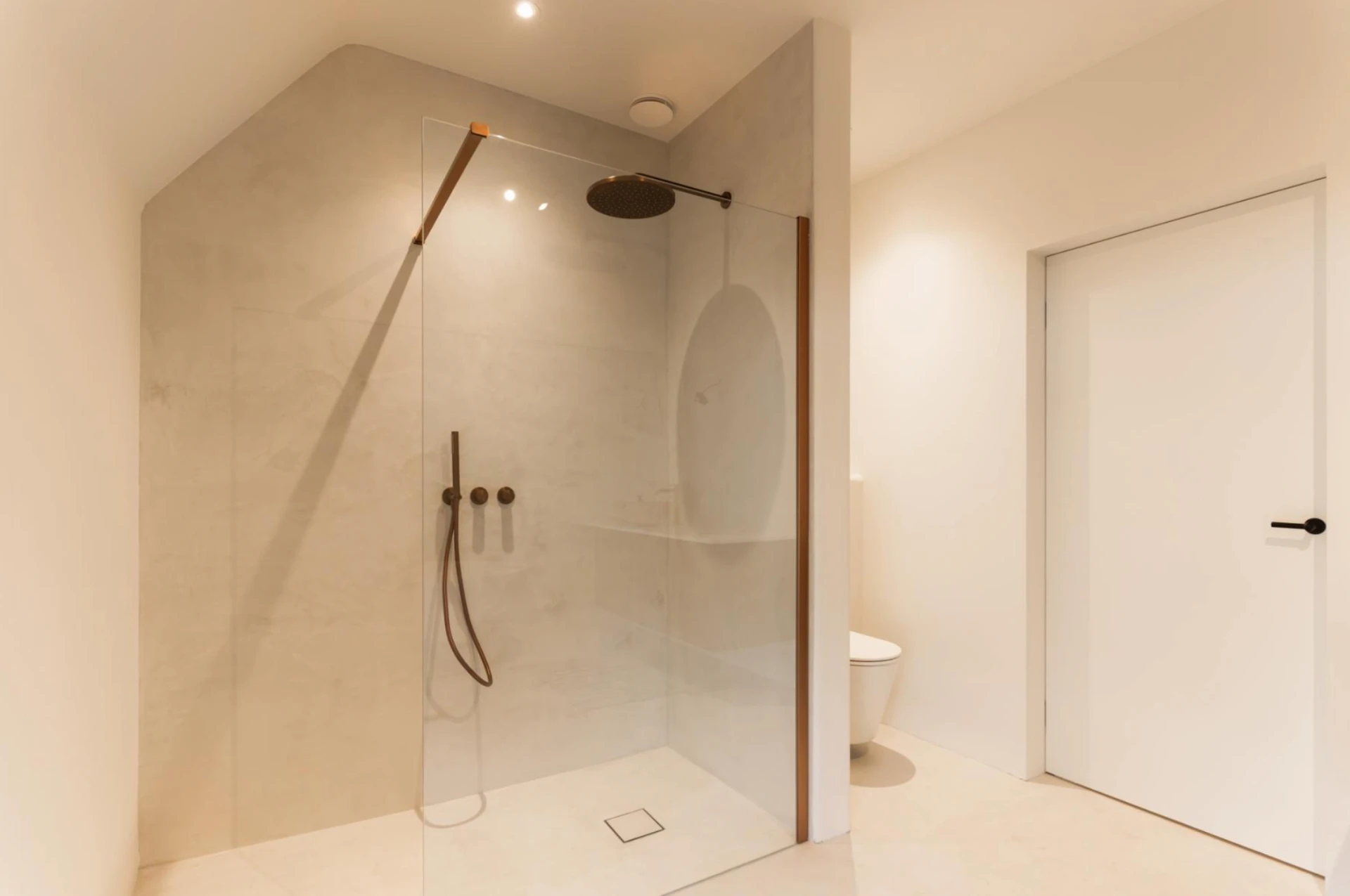 Walk-in shower in sand-toned microcement with dark bronze fixtures and warm recessed lighting